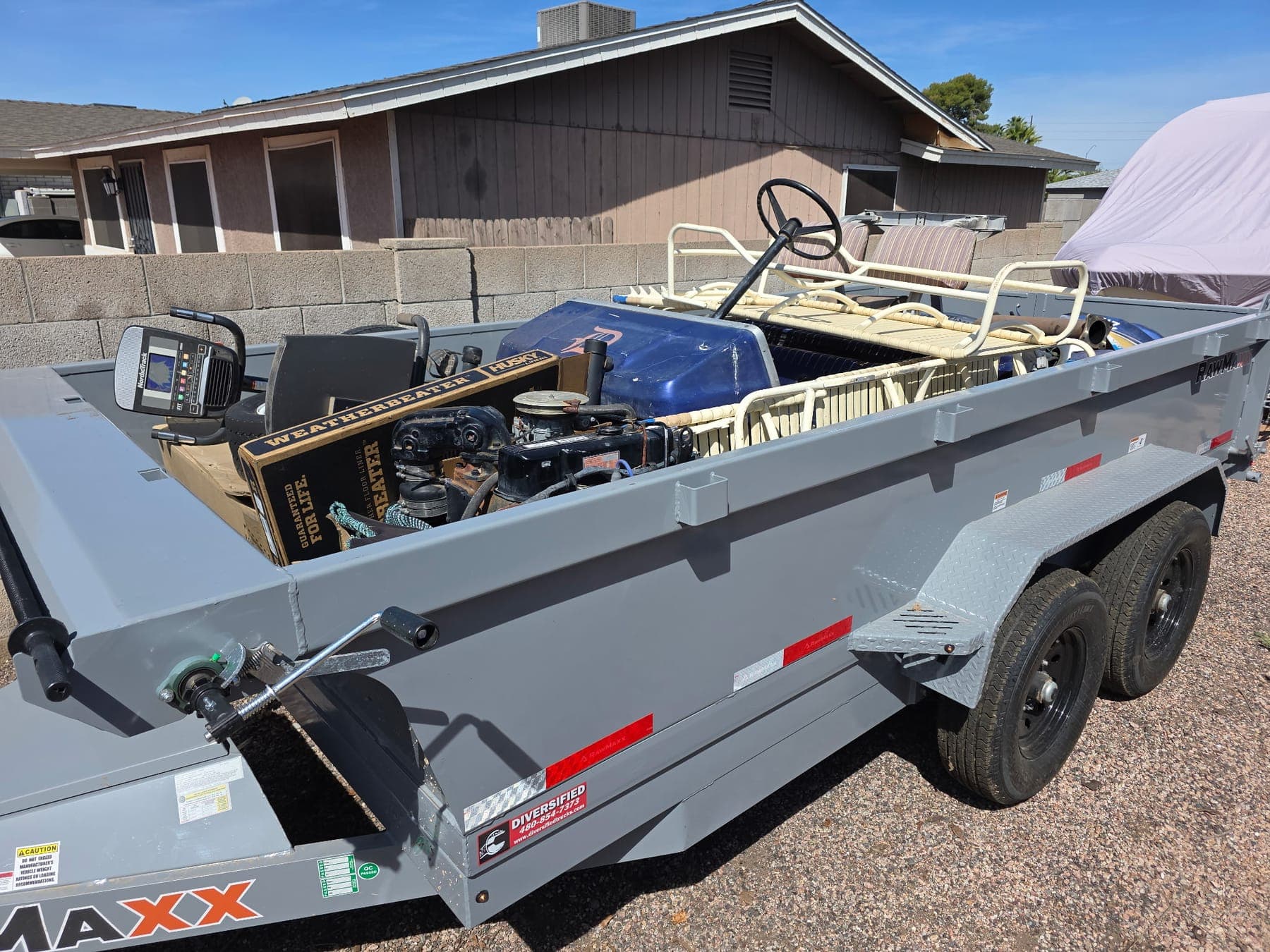 A dump trailer loaded with an old golf cart, an engine, an exercise machine and boxes, parked on a gravel driveway