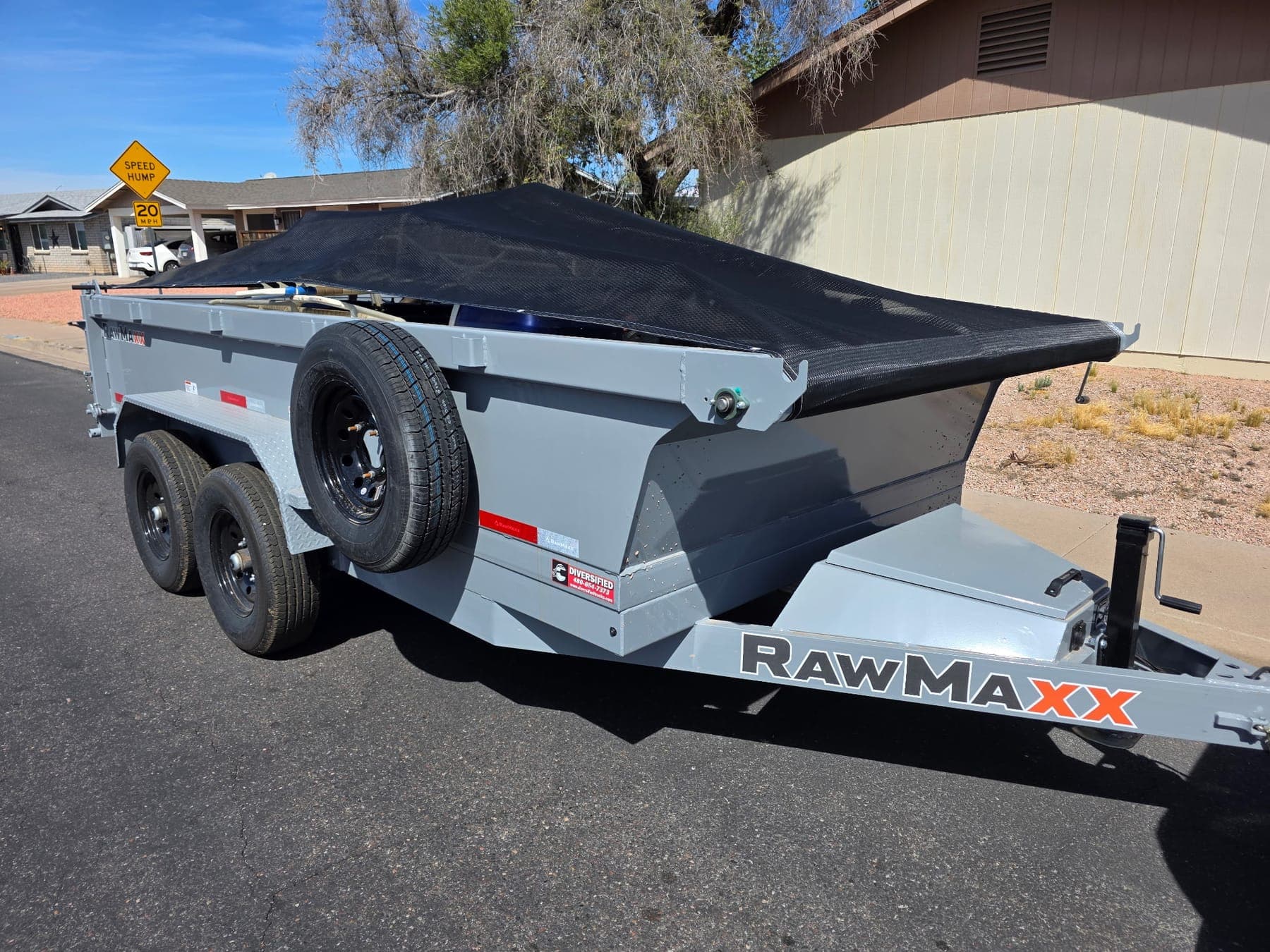 A loaded dump trailer under a black mesh tarp, parked at the curb of a residential street